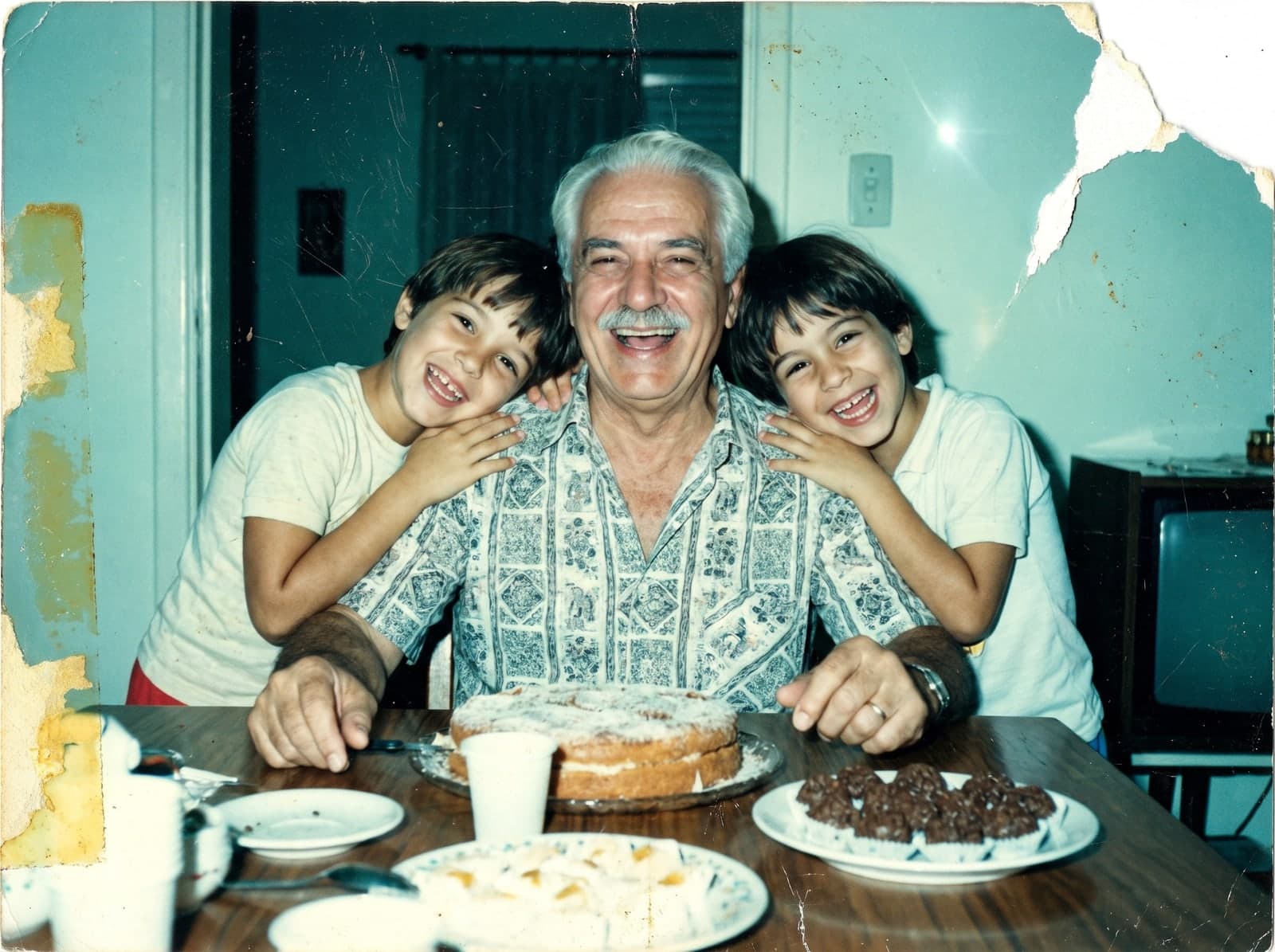Foto antiga de um avô entre dois netos diante de um bolo, com canto rasgado e cor desbotada, antes da restauração