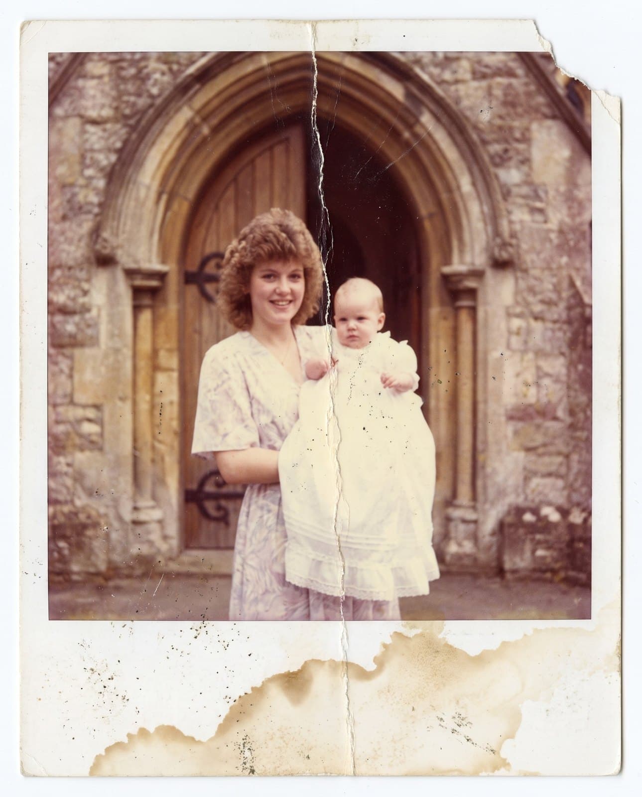 Foto antiga colorida de uma jovem de cabelo cacheado segurando um bebê com vestido branco de batizado diante do portal de uma igreja de pedra, com cor desbotada, vinco no meio, manchas marrons na borda e um canto rasgado