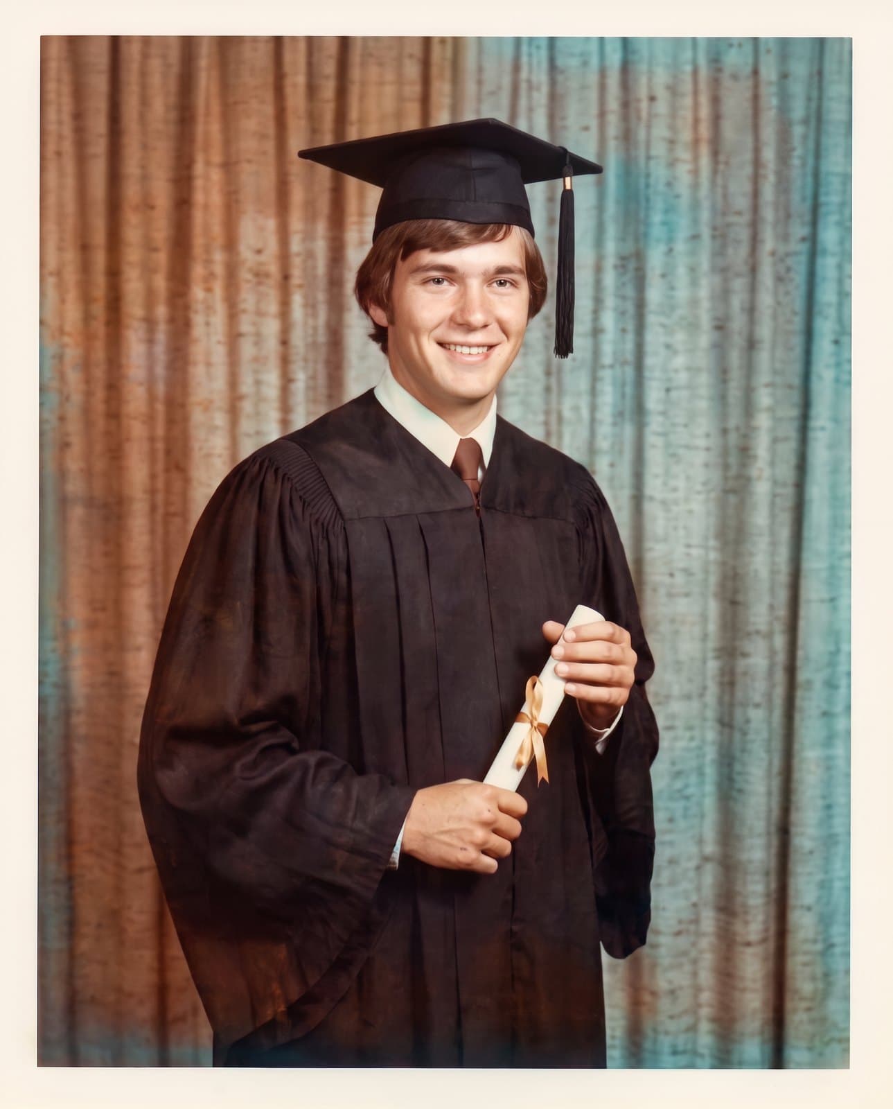 The same photo restored: the young man in cap and gown with the diploma in front of the curtain, in natural colors, with no creases or stains