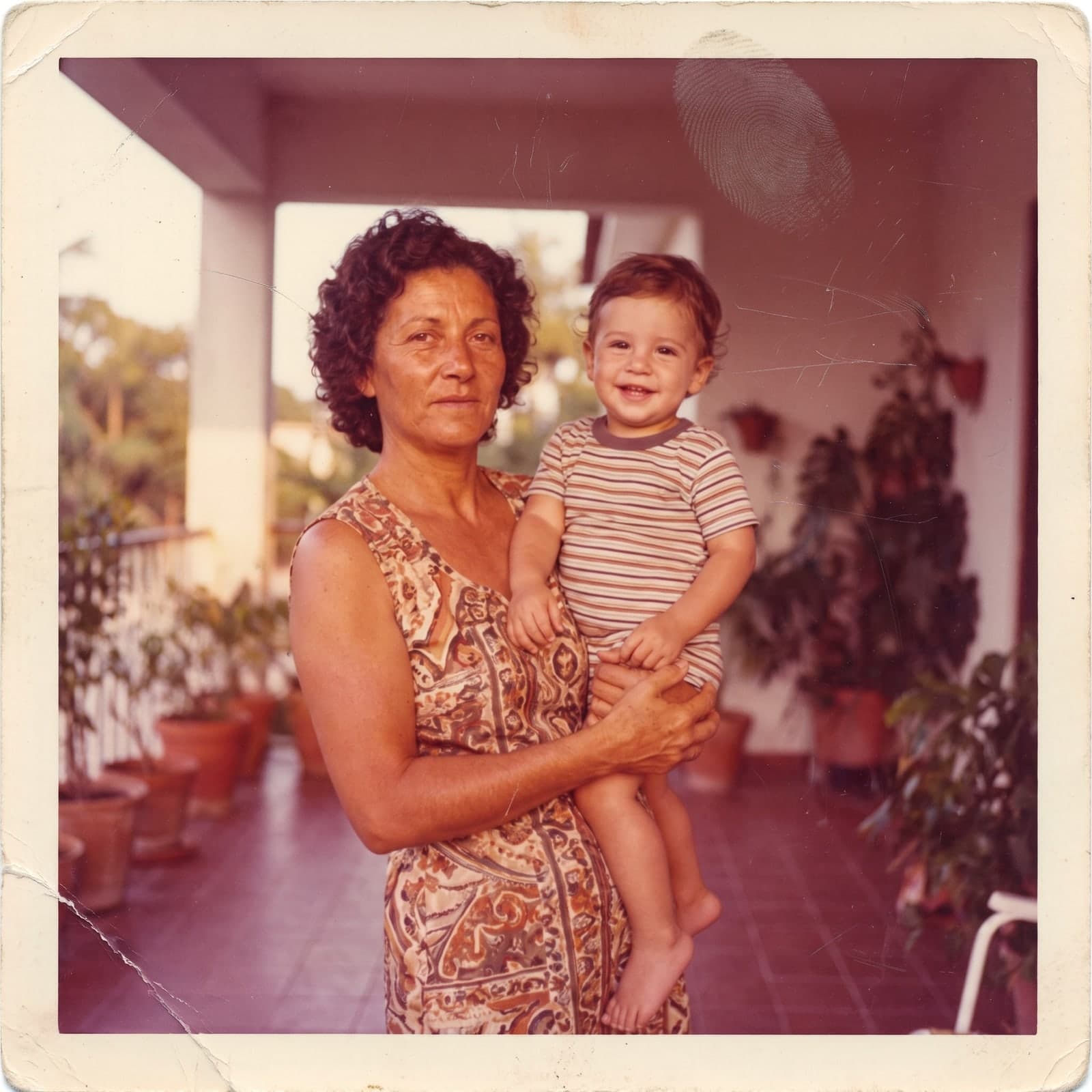 Foto antiga de uma mulher com uma criança no colo numa varanda, com cor desbotada, riscos e uma marca de dedo, antes da restauração
